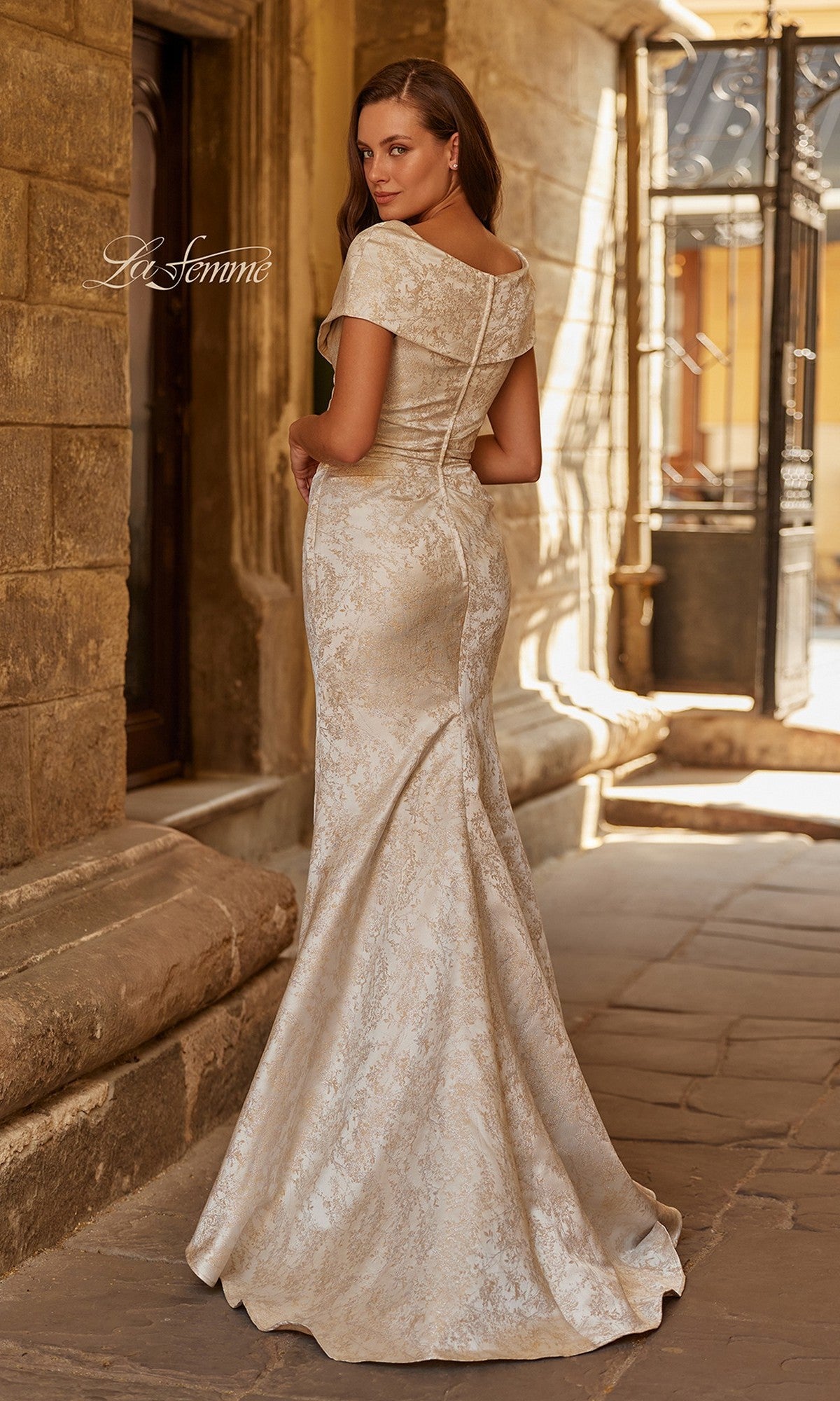 A woman in the La Femme 33428 long formal dress—an off-the-shoulder, beige jacquard evening gown—stands on a stone walkway, glancing over her shoulder at the camera.